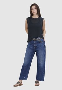 Schwarzes gestricktes ärmelloses Oberteil, blaue locker sitzende Jeans mit fransigem Saum und braune flache Sandalen mit strukturierten Riemen. Verfügt über einen dekorativen Gürtel.