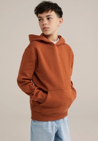 Felpa con cappuccio marrone in tessuto morbido, con tasca a marsupio, polsini a coste e una vestibilità rilassata. Indossata con jeans azzurri chiari.