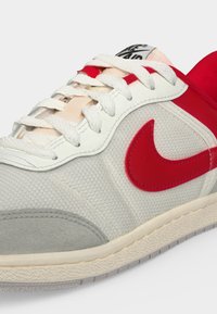 Άσπρο και κόκκινο αθλητικό παπούτσι Nike με λευκά κορδόνια, κόκκινο λογότυπο swoosh, υφασμάτινη υφή και γκρι σουέντ άκρο στα δάχτυλα σε ανοιχτό φόντο.