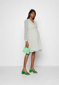 Vestido floral verde claro com mangas longas ajustado na cintura. Combinado com sandálias de salto bloco verdes e uma bolsa a condizer.