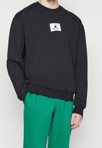 Sudadera negra con cuello redondo y un parche de logo blanco en el pecho, combinada con pantalones de chándal verdes. Tejido suave, corte relajado.