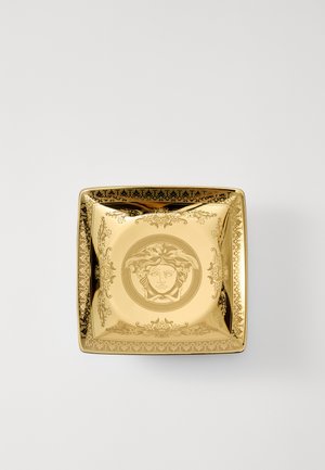 Goldenes quadratisches Schälchen mit kunstvollen schwarzen Details, mittigem Medusa-Motiv und verzierten Rändern. Glatte, reflektierende Oberfläche mit scharfen Kanten.