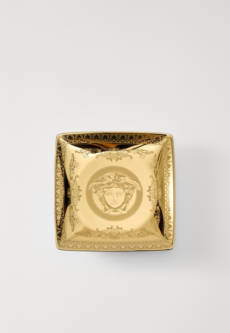 Zelta kvadrātveida bļoda ar smalkām melnām detaļām, centrālais Medūzas motīvs un grezni apmales raksti. Gluda, spīdīga virsma ar asām malām.