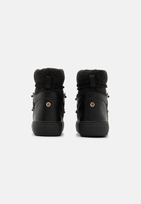 Zapatillas altas negras con un cuello de tela texturizada, suela de goma y detalle de logo dorado en la parte posterior. Cuenta con cordones y una tira de sujeción en bucle.