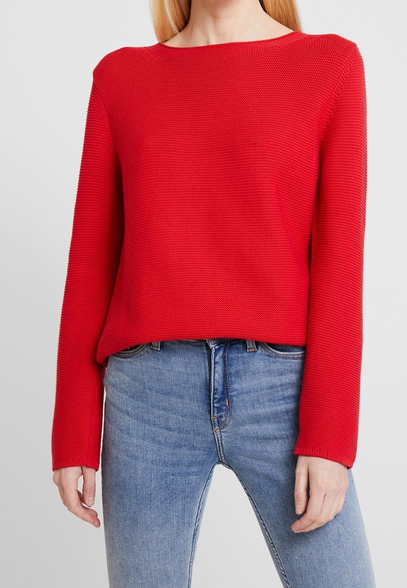 Femme portant un pull en maille rouge à manches longues et un jean bleu, debout devant un fond uni gris clair.