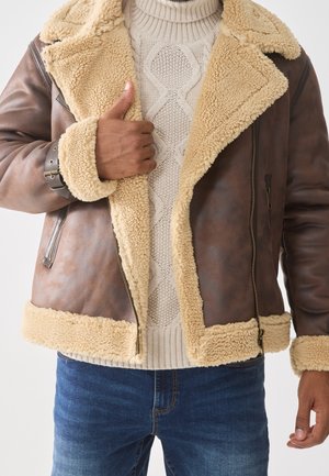 Homme portant une veste en peau retournée marron par-dessus un pull à torsades beige et un jean bleu, avec le pouce levé sur le revers de la veste.