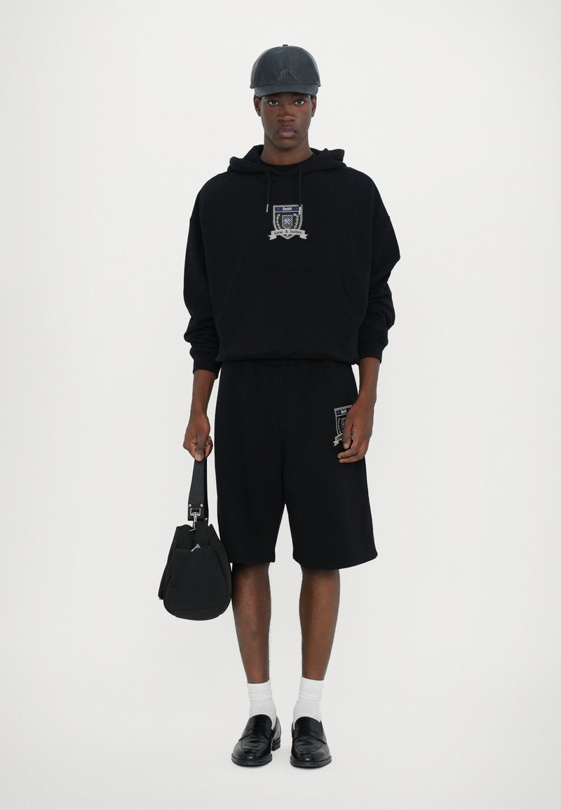 Black hooded sweatshirt with a front pocket and graphic, paired with black shorts, white socks, and black loafers, holding a black bag.