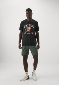 Sort bomulds T-shirt med et farverigt grafisk print med en bulldog og teksten "JUST DO IT," parret med grønne shorts og hvide sneakers.