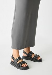Black sandals with two adjustable straps, a thick rubber sole, and metallic buckles. Worn with a long gray garment near the ankle.