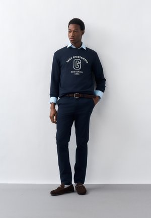 Man wearing navy blue Gant Sportswear sweatshirt over light blue collared shirt, navy trousers, brown belt, and brown loafers, standing against white wall.