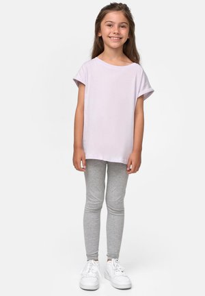 Lächelendes Mädchen mit langem braunen Haar, das ein helllila T-Shirt, graue Leggings und weiße Sneakers trägt, vor einem schlichten Hintergrund stehend.