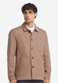 Brun bomullskavaj med krage, två framfickor och rak skärning. Slät struktur och tyg av medelvikts.