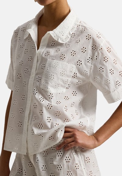 Persoon met een witte korte mouwen blouse met knopen en bijpassende korte broek met oogjes bloemmotieven en borstzak, hand op de heup.