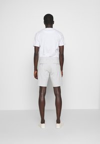 Man standing facing a white wall wearing a white t-shirt, light gray shorts with back pockets, and white sneakers.