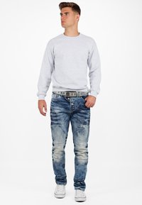 Sweatshirt gris, jeans bleus usés avec des coutures uniques, plusieurs boutons, et une ceinture grise. Des baskets blanches complètent la tenue.
