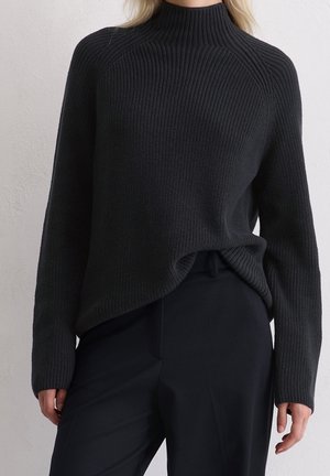 Vrouw draagt een zwart geribd coltrui, ingestopt in zwarte, op maat gemaakte broek, staand tegen een effen lichtgrijze muur.