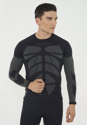Mann trägt schwarzen und grauen Sport-Langarmshirt mit geometrischen Mustern, schaut zur Seite mit Hand an der Hüfte.