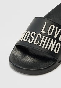 Svart slid sandal med en texturerad läderöverdel och ett brett rem som är präglat med orden "LOVE MOSCHINO" i vitt. Platt sula.