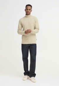 Beige garbó, texturált kötésekkel, sötétkék, egyenes szabású farmernadrággal és fehér sportcipővel párosítva, kezek összekulcsolva elöl.