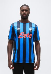 Vyro figūra, stovinti veidu į priekį, dėvinti mėlynos ir juodos spalvų dryžuotą Atalantos futbolo marškinėlį su raudonu „Lete“ logotipu ir juodas kelnes.