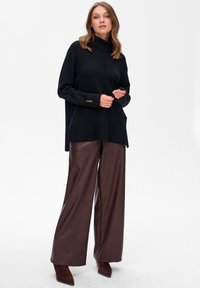 Kobieta w czarnym swetrze z golfem, szerokich brązowych skórzanych spodniach oraz brązowych butach na obcasie ze spiczastym noskiem na jednolitym białym tle.