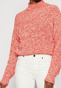 Červený a bílý pletený svetr s vysokým límcem, volný střih a žebrované manžety, v kombinaci s bílými džíny. Texturovaný vzor po celém svršku.