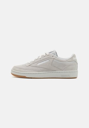 Zapatilla Reebok baja beige de ante con cordones blancos y suela de goma, logo lateral y detalles cosidos sobre un fondo liso.