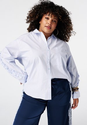 Femme aux cheveux bouclés portant de grandes boucles d’oreilles créoles, une chemise claire rayée boutonnée et un pantalon foncé avec des bandes latérales en dentelle sur fond blanc.