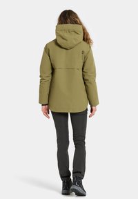 Veste imperméable vert olive avec capuche et ventilation à l'arrière. Associée à un pantalon sombre et des bottes de randonnée noires, mettant en avant un design pratique pour l'extérieur.