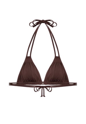 Kobieta o kręconych ciemnych włosach ubrana w brązowy string bikini z topem na szyję i majtkami wiązanymi po bokach na jednolitym jasnym tle.