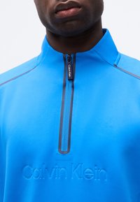 Άνδρας που φοράει φωτεινό μπλε ζακέτα Calvin Klein με ανάγλυφο λογότυπο και μαύρο φερμουάρ, κοντινή λήψη από το στήθος μέχρι το πηγούνι.