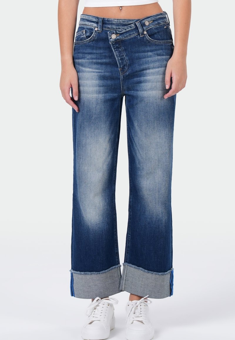 Donkerblauwe wijde jeans met een vervaagde afwerking, voorzien van een omgeslagen zoom in lichte denim. Lage taille met metalen knoopdetails. Witte sneakers.