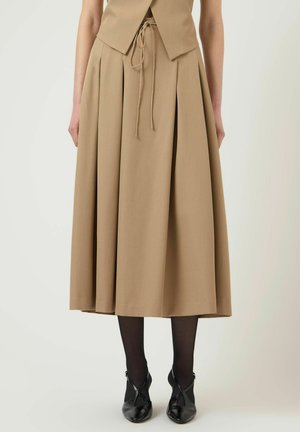 Beige midinederdel med høj talje og forkantplissering, bundet taljesnor, båret med sorte strømpebukser og sorte spidse sko med krydsremme.