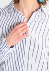 Camicia con bottoni caratterizzata da strisce verticali blu e bianche, con colletto e maniche lunghe. Il tessuto sembra leggero e testurizzato.