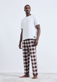 Άνδρας που στέκεται ξυπόλητος, φορώντας λευκό μπλουζάκι κοντό μανίκι και κόκκινα-λευκά καρεδάκια παντελόνια, μπροστά από ένα απλό λευκό φόντο.