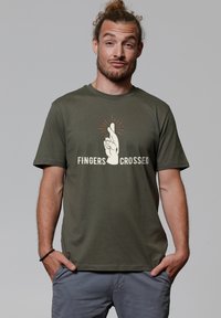 Olivgrünes Baumwoll-T-Shirt mit einem Grafikdesign eines aufgeschlossenen Hand mit gekreuzten Fingern und Strahlen darüber, sowie dem Text "FINGERS CROSSED" darunter.