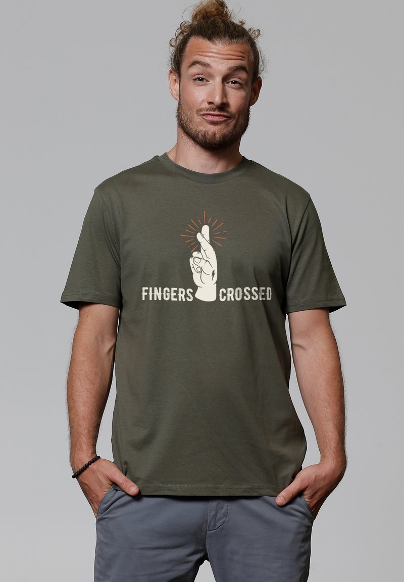 Olivgrünes Baumwoll-T-Shirt mit einem Grafikdesign eines aufgeschlossenen Hand mit gekreuzten Fingern und Strahlen darüber, sowie dem Text "FINGERS CROSSED" darunter.
