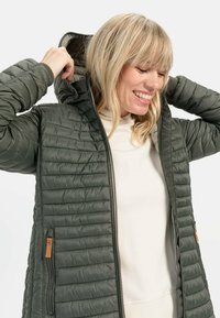 Groene pufferjack met een capuchon, met een gewatteerde textuur, ritssluiting aan de voorkant en zijzakken. Accenten omvatten tan hardware details.