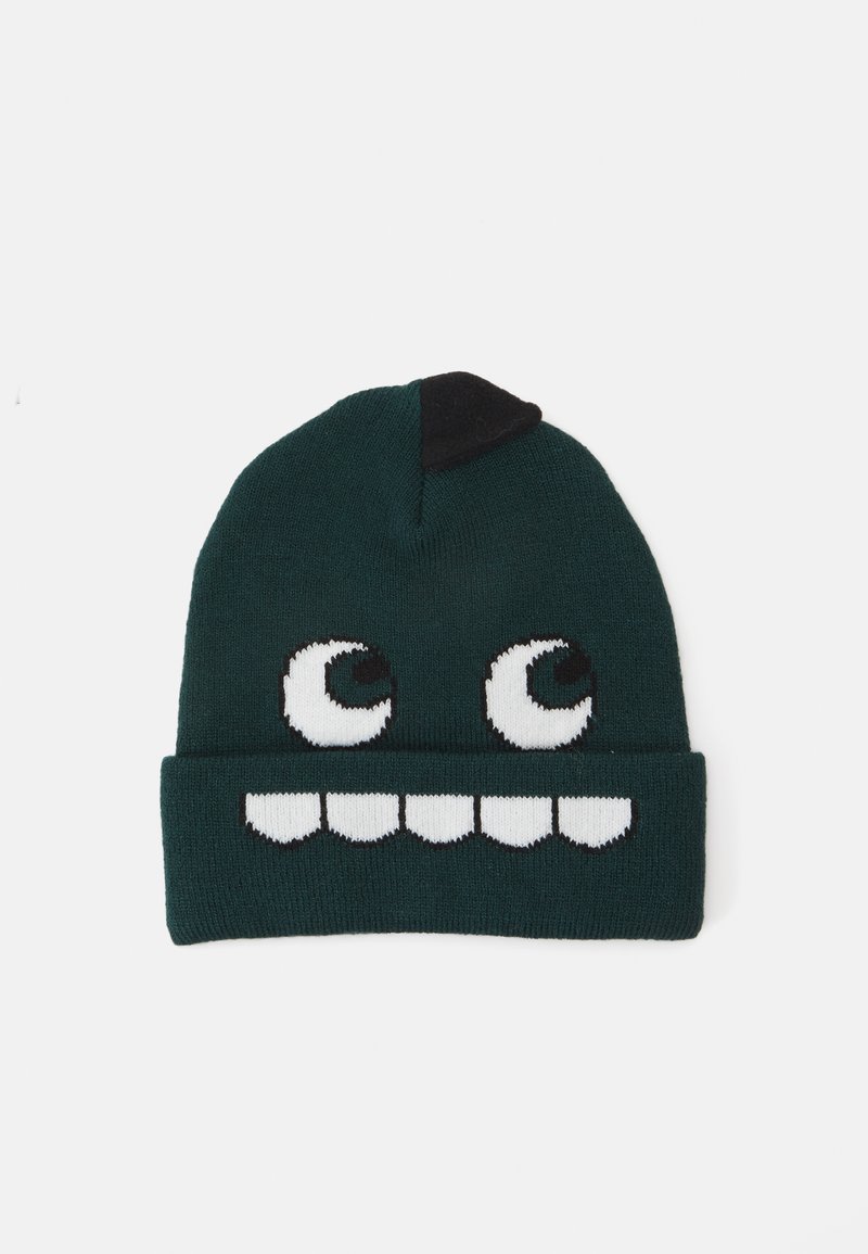Strikket lue i mørkegrønn med svart aksent; har store hvite øyne og et tannete smil mønster. Myk tekstur, uformell design.