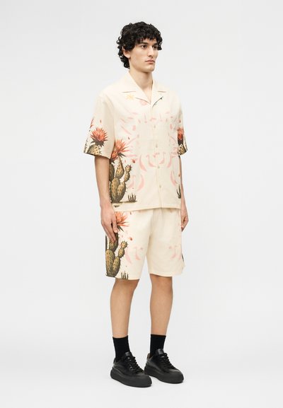 Νεαρός άνδρας που φοράει μπεζ κοντομάνικο πουκάμισο με κουμπιά και ταιριαστά σορτς με σχέδιο κάκτου, μαύρες κάλτσες και μαύρα αθλητικά παπούτσια, στέκεται ακίνητος.