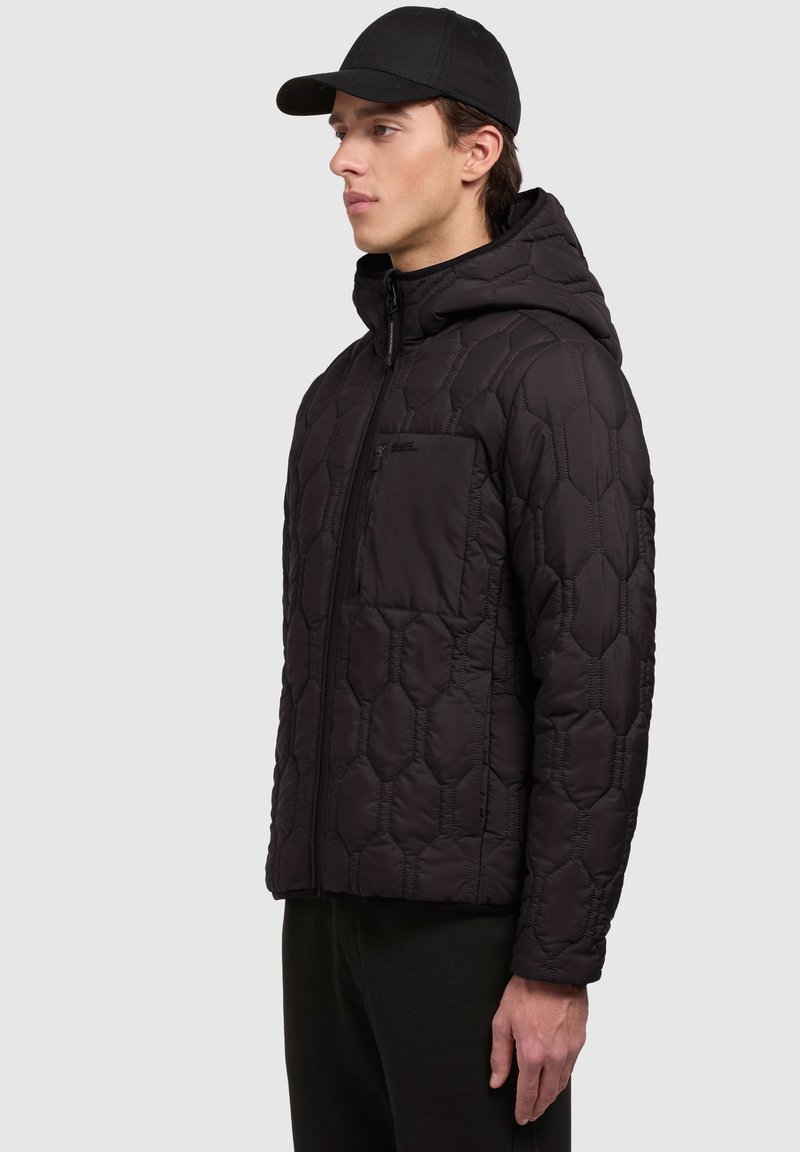 Veste matelassée noire avec capuche, dotée d'une fermeture éclair à l'avant, d'un motif hexagonal et d'une poche poitrine. Matière brillante, portée avec une casquette noire.