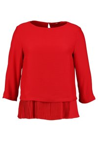 Blouse rouge à manches longues avec un col rond, dotée d'un bas plissé et d'une petite ouverture en forme de clé dans le dos. Tissu au toucher lisse.