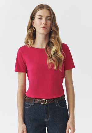Rotes Kurzarmoberteil mit rundem Ausschnitt, kombiniert mit dunkelblauen Jeans und einem braunen geflochtenen Gürtel. Glatte Stoffstruktur.