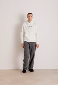 Felpa bianca con cappuccio e logo 'Calvin Klein', abbinata a pantaloni grigi a gamba larga e scarpe nere, su uno sfondo semplice.