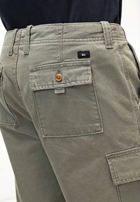 Pantalones cargo de algodón en verde oliva con un bolsillo trasero con tapa y un botón de madera. Incluye una etiqueta negra con el logo cerca de la cinturilla.