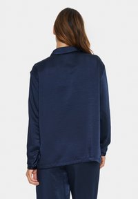 Navy blue satin shirt with long sleeves, a collared neckline, and a smooth texture, featuring a relaxed fit and minimal detailing.