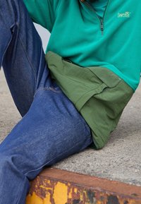 Sudadera de forro polar verde con cremallera, superpuesta sobre una tela verde oliva, combinada con jeans de mezclilla azul oscuro que muestran costuras y textura visibles.