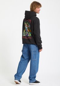 Zwarte hoodie met kleurrijke grafische afbeelding op de achterkant, met tekst. Gecombineerd met losse, blauwe denim broek en zwarte sneakers.