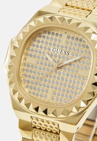Reloj dorado con una correa texturizada y bisel con tachuelas. La esfera presenta una cuadrícula de piedras brillantes y el prominente logotipo de "GUESS".