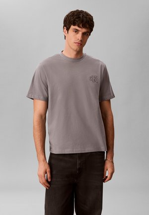 Jeune homme portant un t-shirt gris uni à manches courtes avec un logo "cK" sur la poitrine, associé à un pantalon noir, debout devant un fond uni.